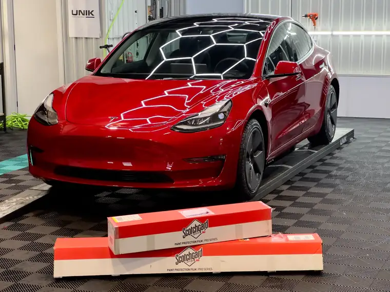 Tesla Model 3 rouge en atelier, devant des boîtes de pellicule pare-pierre 3M Scotchgard (PPF)